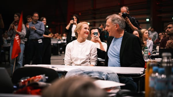 Ines Schwerdtner (l.) & Jan van Aken (r.) sitzen an einem der Tische beim Parteitag und grinsen sich gegenseitig an.